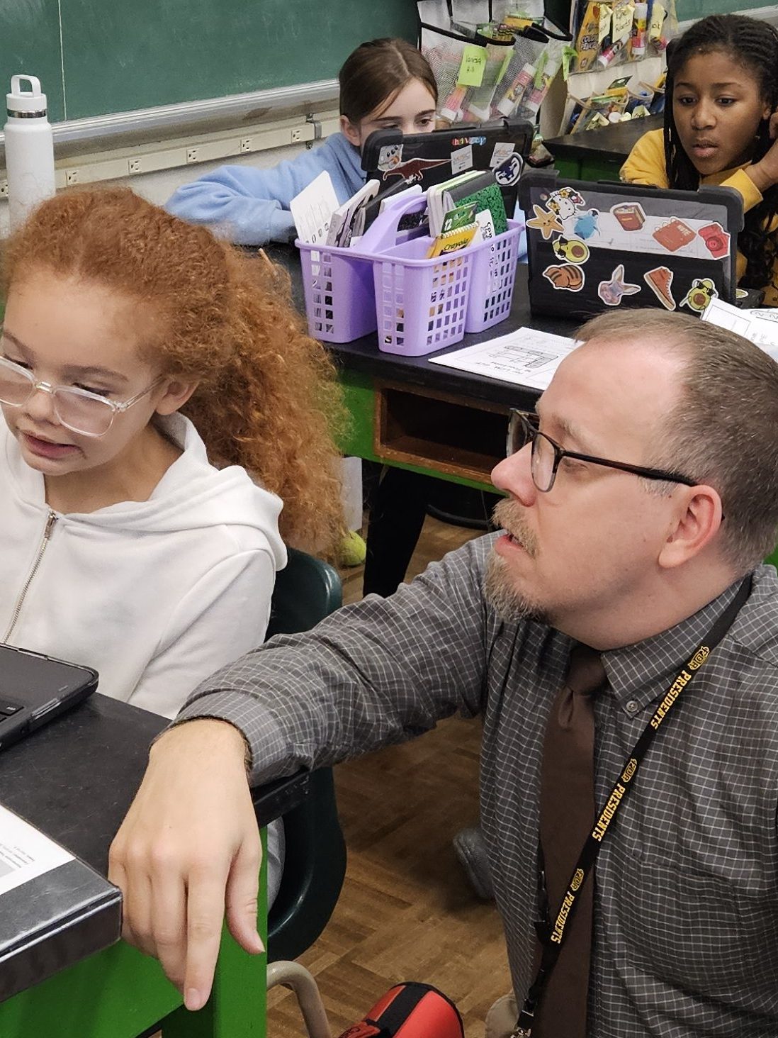David is kneeling down next to s students desk and looking at the computer screen with the student.
