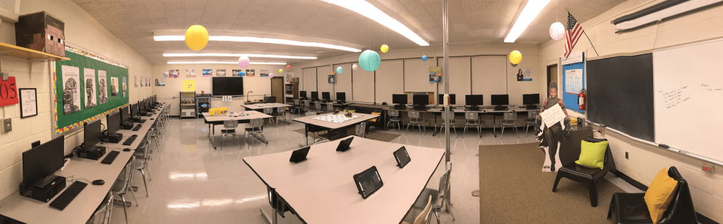 Colorful computer classroom with 
devices around the perimeter and collaborative desks in the middle.